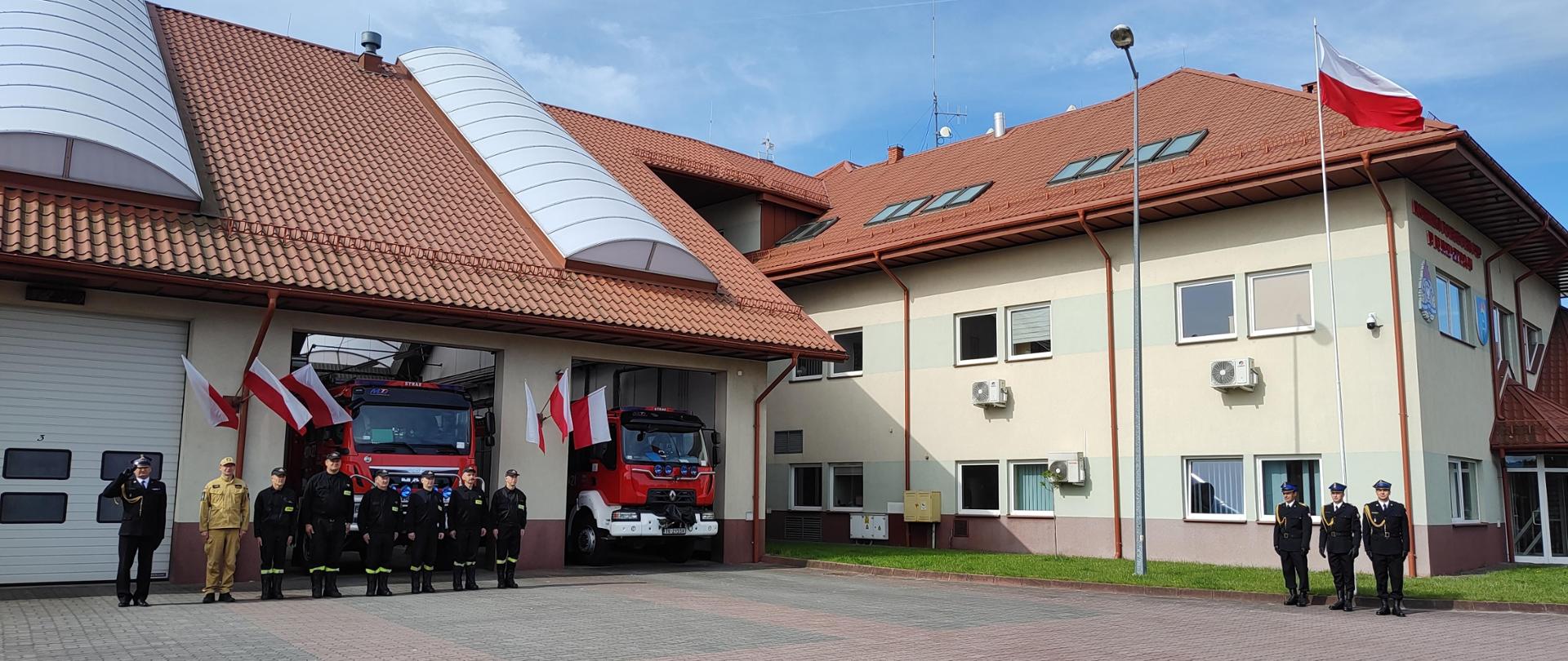 Po prawej stronie stoi 3 strażaków w mundurach galowych. Nad nimi flaga koloru biało czerwonego. Na lewo Strażacy stojący w rzędzie, jeden z nich oddaje honor palcami do daszka czapki. W tle samochody pożarnicze. 