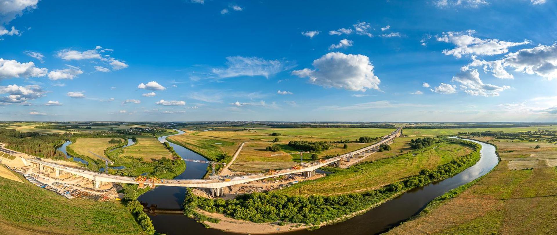Widok z lotu ptaka na wielką plac budowy mostu nad meandrującą rzeką. Rzeka wygina się przez krajobraz, zielone pola i skupiska drzew po obu stronach. Na placu budowy widać wiele żurawi, kilka częściowo ukończonych odcinków mostu z widocznymi rusztowaniami oraz liczne pojazdy budowlane. Niebo jest bezchmurne, co sugeruje dobre warunki pogodowe do prac budowlanych.