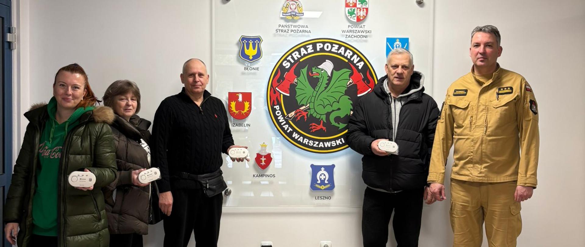 Zdjęcie przedstawia grupę pięciu osób stojących w pomieszczeniu, które wygląda na siedzibę straży pożarnej w Polsce.
Oto szczegółowy opis tego, co widać na fotografii:
Postacie i przedmioty
Grupa osób: W kadrze znajdują się dwie kobiety i trzech mężczyzn. Czworo z nich (dwie kobiety po lewej oraz dwaj mężczyźni w centrum) trzyma w dłoniach małe, białe urządzenia elektroniczne – prawdopodobnie czujniki tlenku węgla (czadu) lub dymu.
Strażak: Po prawej stronie stoi mężczyzna w piaskowym mundurze służbowym Państwowej Straży Pożarnej, na którym widoczne są pagony ze stopniem starszego brygadiera (trzy gwiazdki i dwa paski).
Ubiór pozostałych osób: Pozostali uczestnicy są ubrani w stroje cywilne, kurtki zimowe i bluzy, co sugeruje, że mogą to być mieszkańcy lub przedstawiciele lokalnej społeczności odbierający urządzenia.
Tło i symbolika
Tablica informacyjna: W centralnym punkcie, na ścianie, wisi duża przezroczysta tablica z logotypami. Główny logotyp to godło Straży Pożarnej Powiatu Warszawskiego Zachodniego z charakterystycznym zielonym smokiem.
Herby: Wokół centralnego logo umieszczone są herby gmin wchodzących w skład powiatu, m.in. Błonia, Izabelina, Kampinosu i Leszna. Na górze widoczne jest godło Państwowej Straży Pożarnej oraz herb Powiatu Warszawskiego Zachodniego.
Otoczenie: Pomieszczenie jest jasne, o surowym, urzędowym charakterze, z kafelkami na podłodze i kasetonowym sufitem.