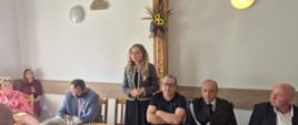 Spotkanie uczestników dożynek w sali. Wojewódzka Inspektor stoi przed grupą osób siedzących przy stole; wśród nich znajduje się osoba w mundurze strażackim. W tle widoczna dekoracja z suszonych roślin i słoneczników.