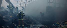 Strażacy w akcji gaszenia pożaru na składowisku odpadów. Widoczny jest szkielet wiaty, otoczony gęstym dymem. Dwóch strażaków z wężami gaśniczymi walczy z ogniem, a w tle widać koparkę pomagającą w rozbiórce lub rozgarnianiu palących się materiałów. Na pierwszym planie leżą rozrzucone śmieci.