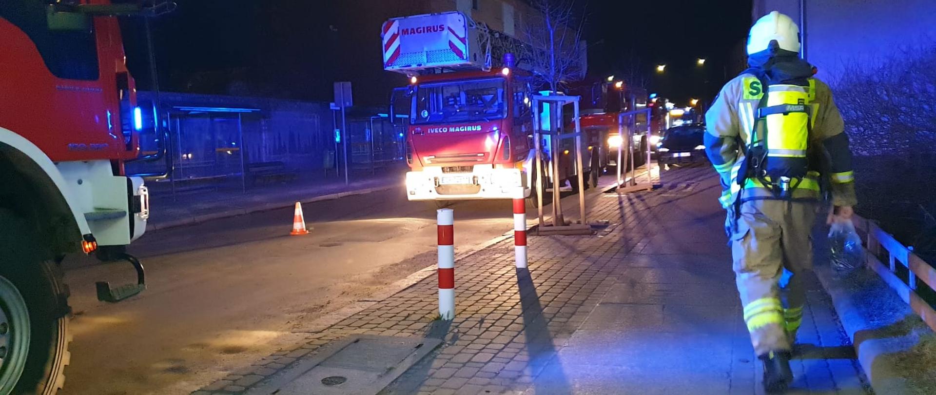 Na zdjęciu widoczne samochody pożarnicze koloru czerwonego, stojące na jezdni przy chodniku. Zdjęcie wykonane podczas działań ratowniczo-gaśniczych przy pożarze na ulicy Duboisa w Opolu. Po prawej stronie widoczny strażak OSP idący w stronę pojazdów (odwrócony plecami) w ubraniu specjalnym koloru piaskowego, białym hełmie na głowie oraz aparacie powietrznym na plecach. 