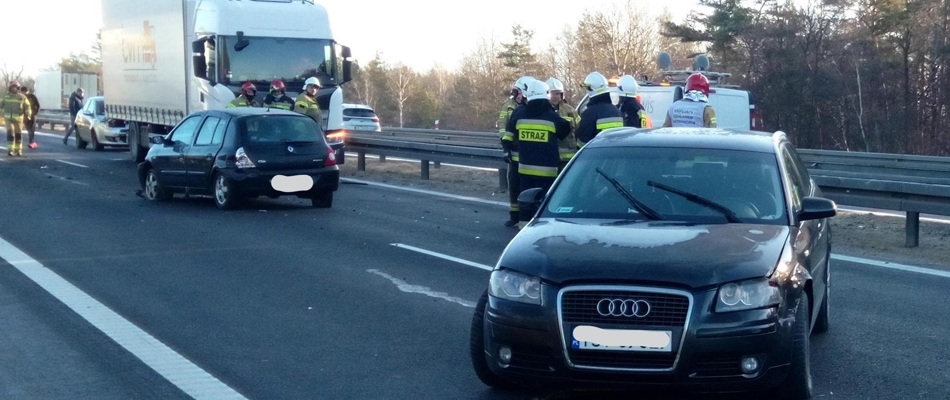 Na zdjęciu przedstawiono perspektywę drogi S7. Na pierwszym planie stoi samochód osobowy Audi a nieco dalej Renault –auta po kolizji. Miedzy pojazdami grupa strażaków. W dalszej perspektywie samochód ciężarowy i kolejna grupa strażaków.