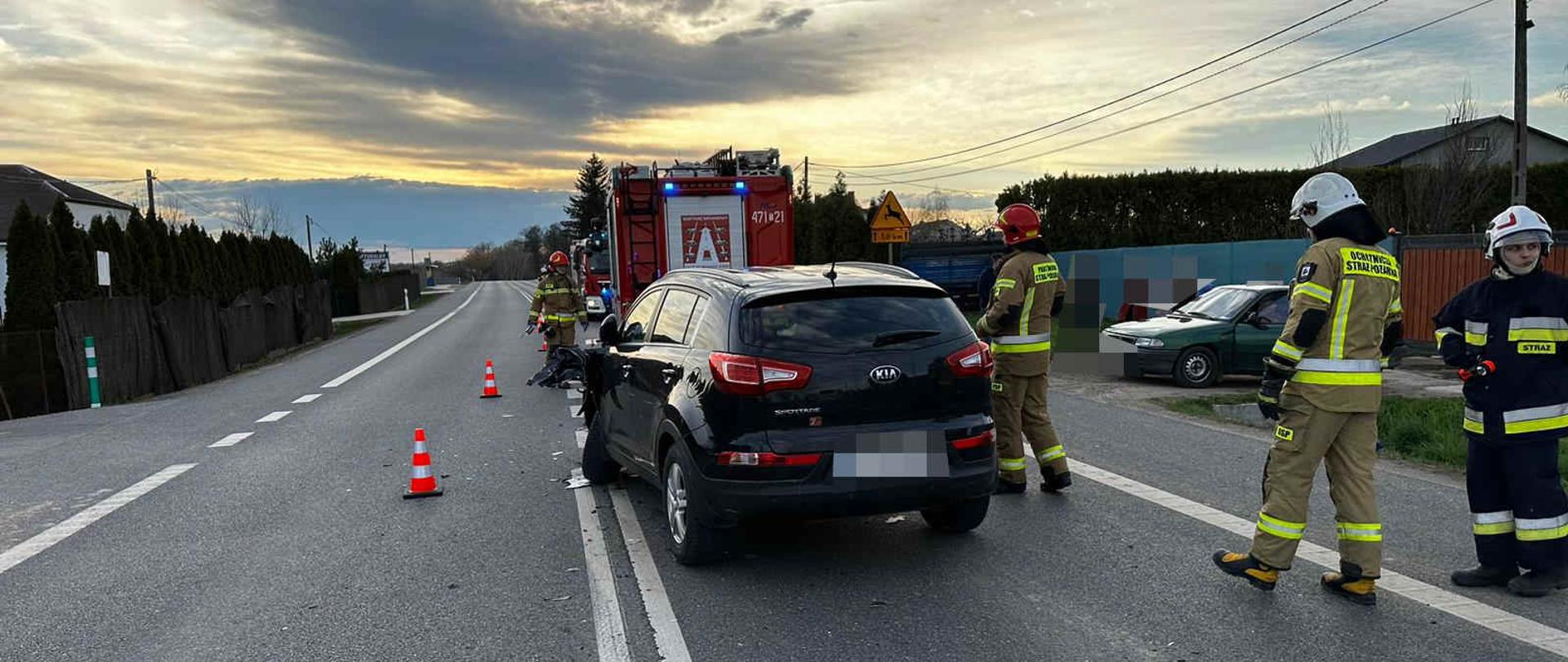 Zdjęcie – przedstawia samochód biorący udział w wypadku oraz strażaków pracujących na miejscu zdarzenia. Na drugim planie widać samochód pożarniczy. 