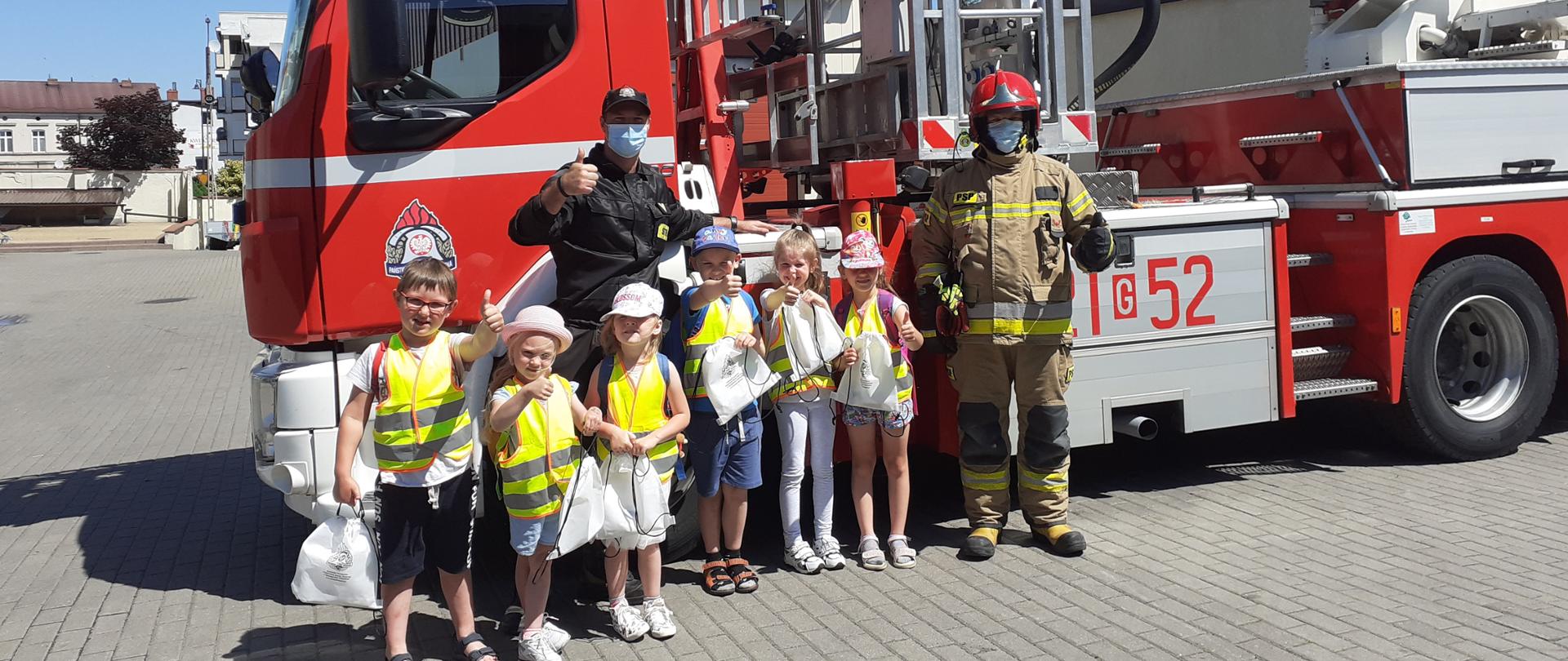 Zdjęcie przedstawia uczestników zajęć w sali ognik na tle podnośnika Komendy, dzieci pokazują zadowolenie gestem uniesionego kciuka