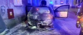 Zdjęcie wykonane w nocy. W centralnym punkcie zdjęcia spalony samochód osobowy VW GOLF 4 stojący przy kamienicy i widoczny od tyłu. Zdjęcie oświetlają niebieski lampy błyskowe z samochodu gaśniczego