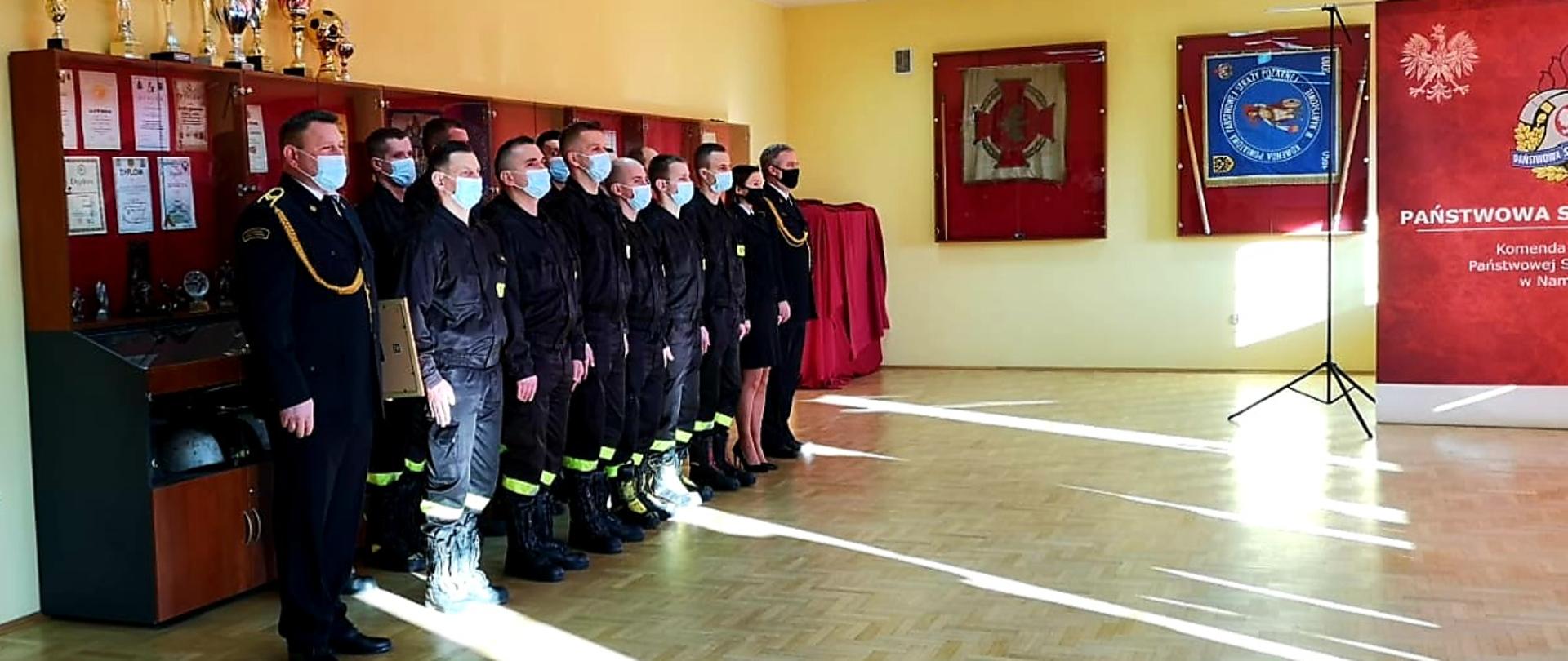 Strażacy w dwuszeregu podczas uroczystej zbiórki. Pierwszy od lewej strażak w mundurze wyjściowym, następnie część pododdziału w umundurowaniu koszarowym. Dwoje ostatnich funkcjonariuszy w umundurowaniu wyjściowym.