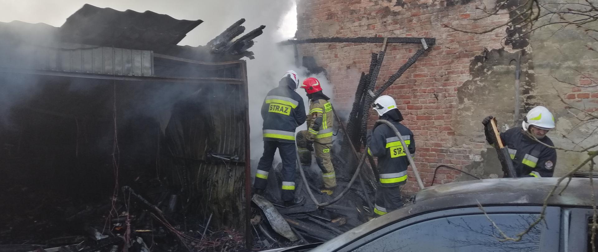 Na środku zdjęcia widoczni są czterej strażacy ubrani w nomexy oraz hełmy, którzy dogaszają i przelewają garaż który uległ spaleniu w wyniku pożaru oraz wynoszą spalone drewniane elementy. Po prawej stronie zdjęcia widoczny jest murowany budynek wykonany z czerwonej cegły , do którego przylegał spalony garaż. Po lewej stronie stoi blaszany garaż, który również uległ zniszczeniu w wyniku pożaru. Posiada powyginaną i zdeformowaną blachę koloru szarego. Nad garażem unosi się chmura siwego dymu. W prawym dolnym rogu na zdjęciu widać również część samochodu osobowego koloru srebrnego, u którego w wyniku pożaru spaleniu uległa przednia maska oraz dach samochodu. Nad samochodem znajdują się odstające gałęzie.