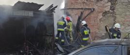  Na środku zdjęcia widoczni są czterej strażacy ubrani w nomexy oraz hełmy, którzy dogaszają i przelewają garaż który uległ spaleniu w wyniku pożaru oraz wynoszą spalone drewniane elementy. Po prawej stronie zdjęcia widoczny jest murowany budynek wykonany z czerwonej cegły , do którego przylegał spalony garaż. Po lewej stronie stoi blaszany garaż, który również uległ zniszczeniu w wyniku pożaru. Posiada powyginaną i zdeformowaną blachę koloru szarego. Nad garażem unosi się chmura siwego dymu. W prawym dolnym rogu na zdjęciu widać również część samochodu osobowego koloru srebrnego, u którego w wyniku pożaru spaleniu uległa przednia maska oraz dach samochodu. Nad samochodem znajdują się odstające gałęzie.