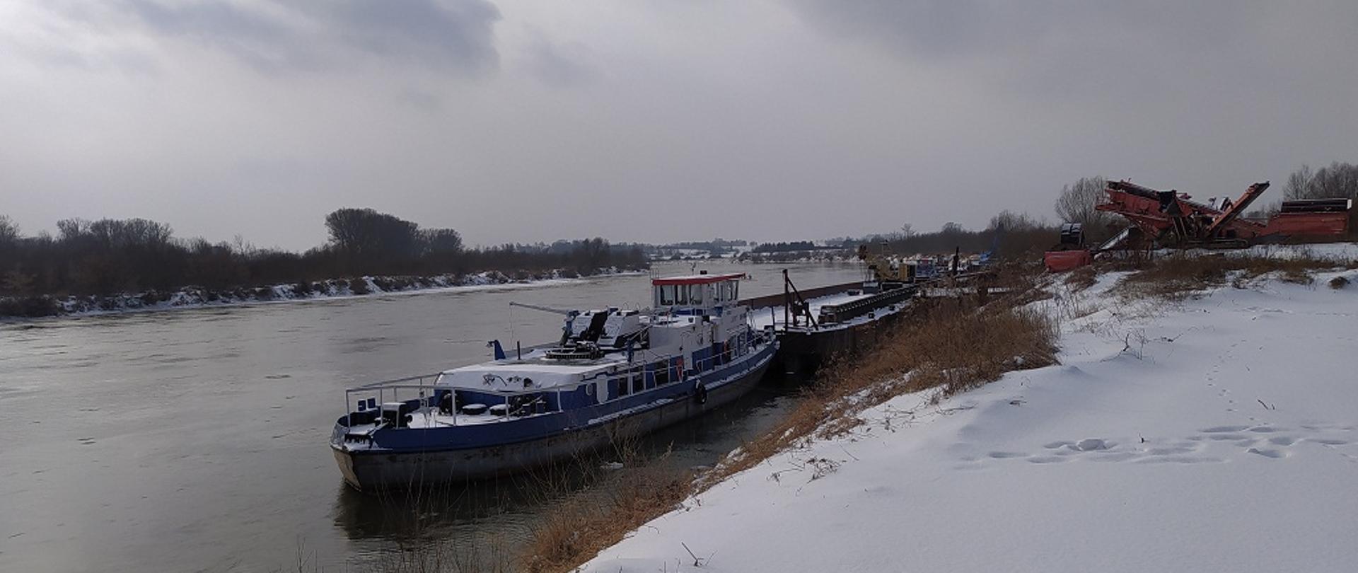 1. Fragment koryta rzeki Wisły z zaśnieżonym brzegiem, na której z prawej strony przy zacumowana barka.