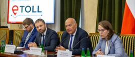Stół prezydialny przy którym siedzą minister finansów Tadeusz Kościński, szefowa KAS Magdalena Rzeczkowska, pełnomocnik ministra finansów ds. informatyzacji Przemysław Koch, w tle slajd prezentacji z napisem e-Toll.