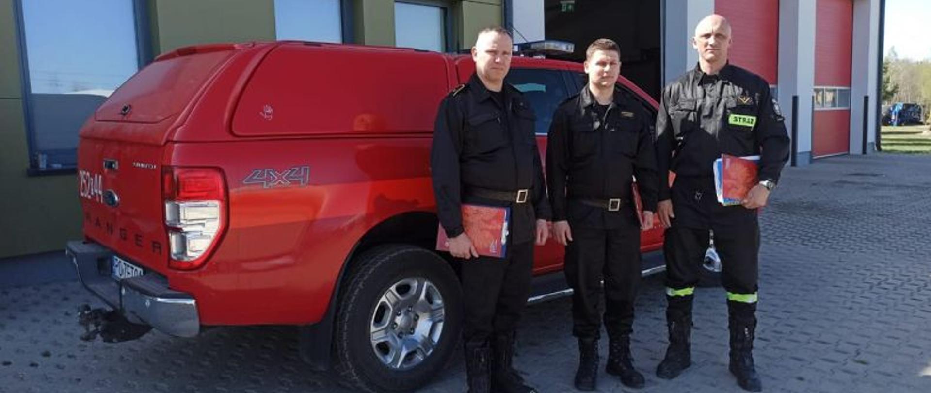 Centralny punkt zdjęcia stanowią trzej strażacy. W tle auto terenowe, czerwone strażackie. Strażacy zadowoleni pozują po ukończeniu szkolenia.