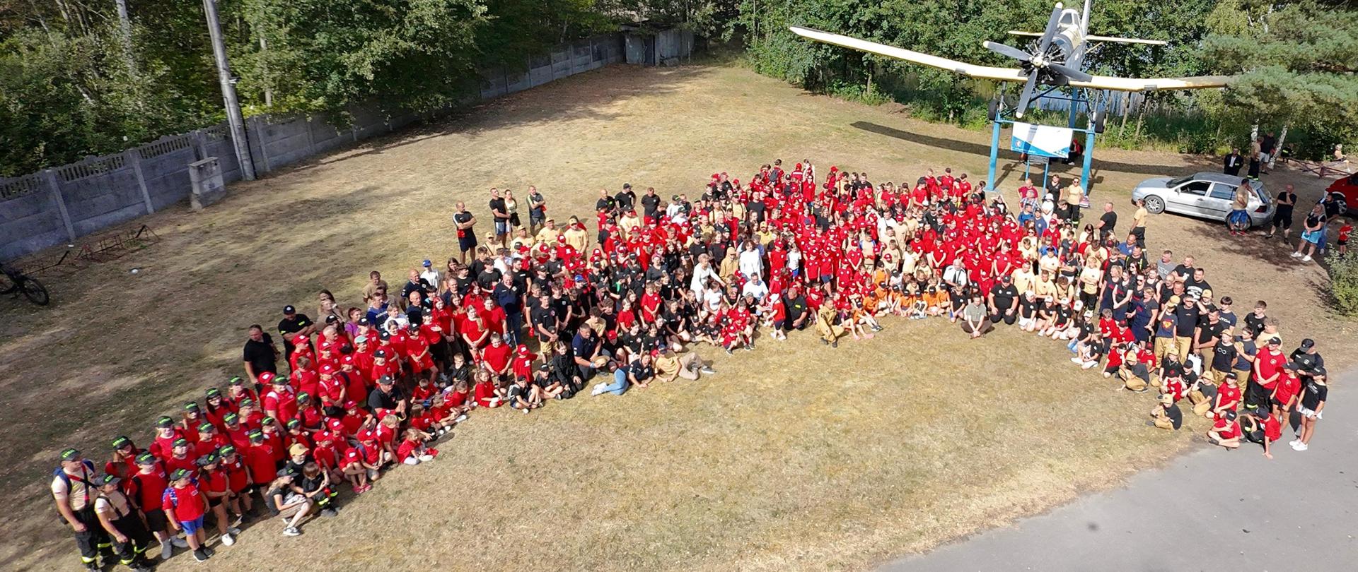 Bardzo duża, kilkusetosobowa grupa uczestników wydarzenia ustawiona jest na placu pod żółto-białym samolotem na stojaku. Na pierwszym planie dominują czerwone koszulki MDP, czarne koszulki OSP oraz beżowe mundury PSP. Widać dzieci, młodzież i dorosłych strażaków. Zdjęcie wykonane zostało z drona, obejmuje całą grupę, fragment drogi oraz drzewa w tle.