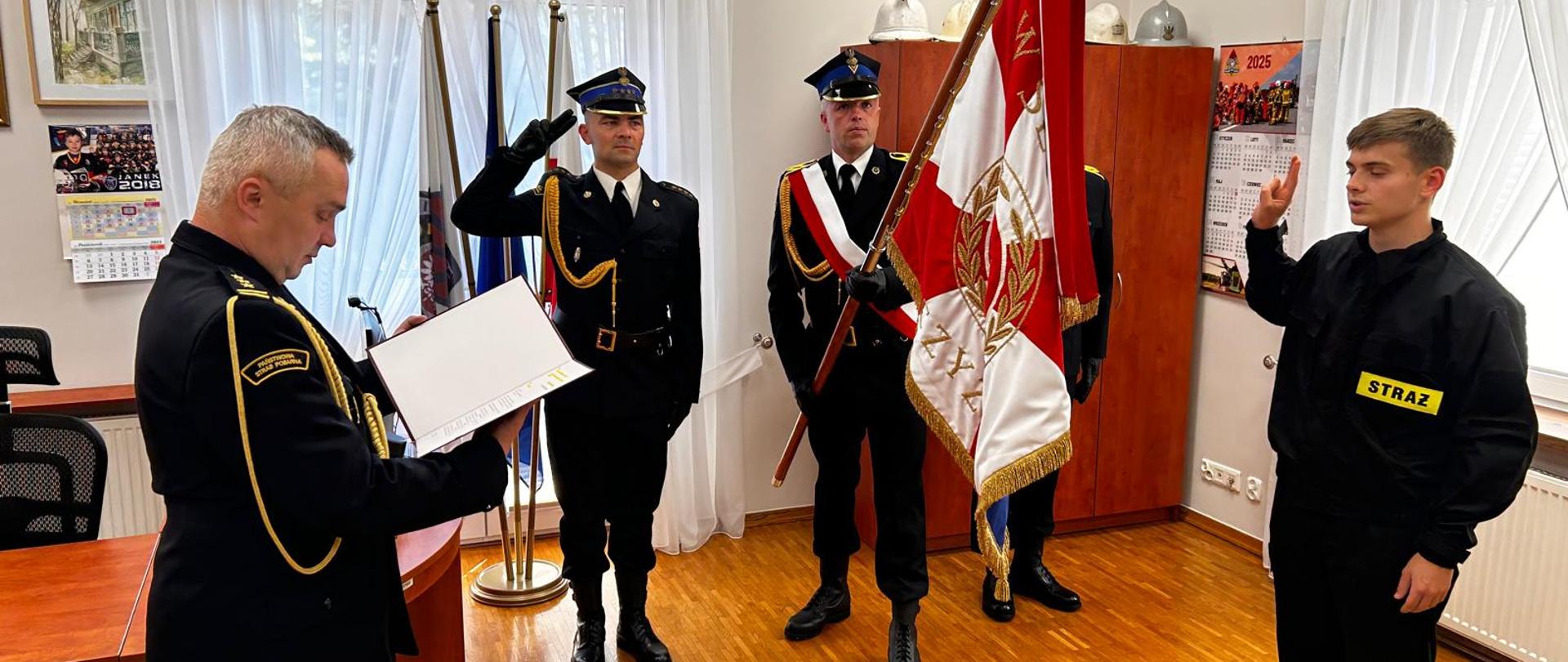 Na zdjęciu widoczna jest uroczystość ślubowania lub zaprzysiężenia strażaka w pomieszczeniu biurowym, najprawdopodobniej należącym do jednostki straży pożarnej w Polsce.
Opis postaci i sytuacji:
Po lewej stronie znajduje się starszy mężczyzna w galowym mundurze strażackim. Trzyma otwarty dokument — prawdopodobnie tekst roty ślubowania — i odczytuje go. Na jego mundurze widnieje naszywka „Państwowa Straż Pożarna”.
Pośrodku stoją trzej funkcjonariusze w galowych mundurach. Jeden z nich trzyma sztandar z polskimi barwami narodowymi oraz symbolem straży pożarnej. Dwóch pozostałych salutuje.
Po prawej stronie stoi młody mężczyzna w mundurze bojowym z wyraźnym napisem „STRAŻ” — najprawdopodobniej nowo przyjęty strażak. Unosi prawą rękę w geście przysięgi, co sugeruje, że składa ślubowanie na sztandar.
Tło i otoczenie:
Pomieszczenie wygląda na urząd lub biuro komendanta. Na ścianach widnieją:
kalendarze strażackie (w tym z rokiem 2025),
obrazy i pamiątki,
kilka hełmów strażackich ustawionych na szafie,
godło Polski (widoczne na innym zdjęciu),
flagi w tle, w tym biało-czerwona oraz strażacka.
Podsumowanie:
Zdjęcie przedstawia podniosły moment ślubowania nowego strażaka w obecności oficjalnego pocztu sztandarowego i przełożonego. Całość ma charakter formalny i ceremonialny, zgodny z tradycją Państwowej Straży Pożarnej w Polsce.
