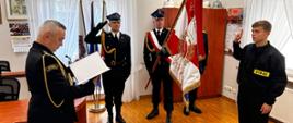 Na zdjęciu widoczna jest uroczystość ślubowania lub zaprzysiężenia strażaka w pomieszczeniu biurowym, najprawdopodobniej należącym do jednostki straży pożarnej w Polsce.
Opis postaci i sytuacji:
• Po lewej stronie znajduje się starszy mężczyzna w galowym mundurze strażackim. Trzyma otwarty dokument — prawdopodobnie tekst roty ślubowania — i odczytuje go. Na jego mundurze widnieje naszywka „Państwowa Straż Pożarna”.
• Pośrodku stoją trzej funkcjonariusze w galowych mundurach. Jeden z nich trzyma sztandar z polskimi barwami narodowymi oraz symbolem straży pożarnej. Dwóch pozostałych salutuje.
• Po prawej stronie stoi młody mężczyzna w mundurze bojowym z wyraźnym napisem „STRAŻ” — najprawdopodobniej nowo przyjęty strażak. Unosi prawą rękę w geście przysięgi, co sugeruje, że składa ślubowanie na sztandar.
Tło i otoczenie:
Pomieszczenie wygląda na urząd lub biuro komendanta. Na ścianach widnieją:
• kalendarze strażackie (w tym z rokiem 2025),
• obrazy i pamiątki,
• kilka hełmów strażackich ustawionych na szafie,
• godło Polski (widoczne na innym zdjęciu),
• flagi w tle, w tym biało-czerwona oraz strażacka.
Podsumowanie:
Zdjęcie przedstawia podniosły moment ślubowania nowego strażaka w obecności oficjalnego pocztu sztandarowego i przełożonego. Całość ma charakter formalny i ceremonialny, zgodny z tradycją Państwowej Straży Pożarnej w Polsce.
