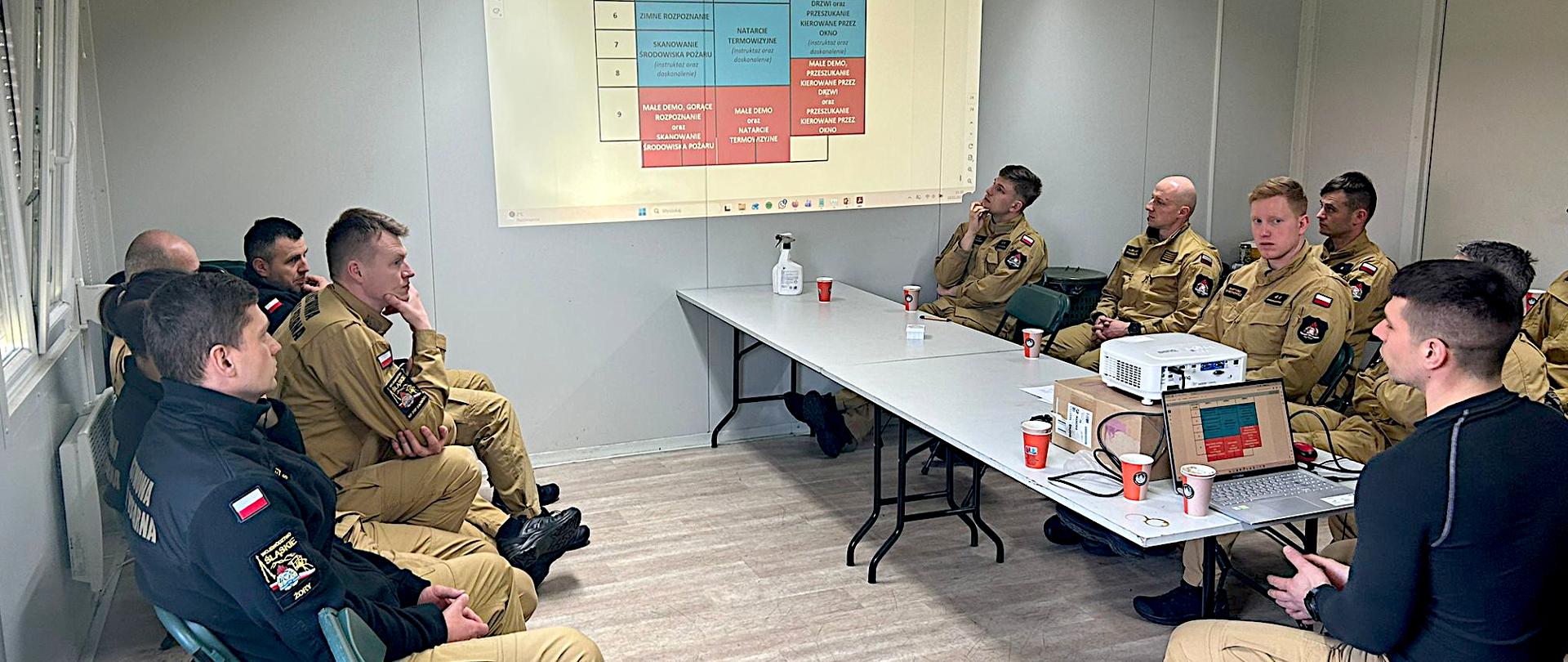 Na zdjęciu widoczna jest grupa strażaków podczas zajęć szkoleniowych w niewielkiej sali. Pomieszczenie jest jasne. Strażacy siedzą na krzesłach ustawionych w kształt litery „U” wokół dwóch stołów. Wszyscy uczestnicy mają na sobie beżowe mundury strażackie z naszywkami – na ramionach widać polską flagę i oznaczenia jednostki. Siedzą w skupieniu i zwróceni w stronę ściany, na której wyświetlany jest obraz z projektora. Po prawej stronie, przy stole, siedzi prowadzący szkolenie – ubrany w czarną koszulkę. Jest odwrócony bokiem do grupy i omawia prezentację. Przed nim stoi laptop podłączony do projektora, a obok leży niewielki projektor wyświetlający obraz na ścianę. Na stołach stoją papierowe kubki.