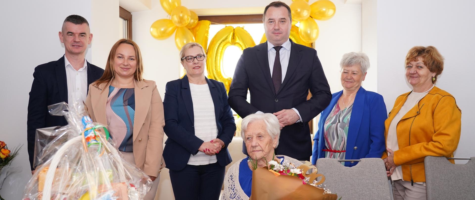 Starsza kobieta z bukietem kwiatów siedzi przy stole, na którym stoi kosz z upominkami za nią stoją cztery kobiety i dwóch mężczyzn. Wszyscy z uśmiechem pozują do zdjęcia.