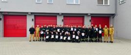 Grupowe zdjęcie strażaków wykonane przed budynkiem remizy strażackiej z trzema czerwonymi bramami garażowymi. Na zdjęciu stoi kilkadziesiąt osób w umundurowaniu strażackim, część z nich trzyma w rękach dyplomy. Budynek ma szarą elewację i dwa piętra. Zdjęcie wykonano na placu z kostki brukowej.