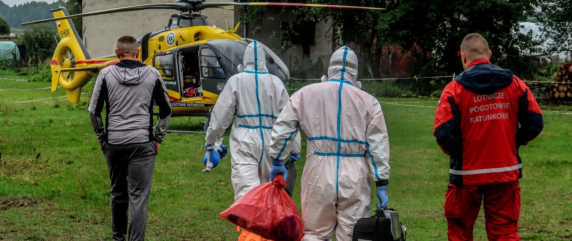 Ubrani w kombinezony ochronni ratownicy medyczni, członek zespołu Lotniczego Pogotowia Ratunkowego oraz osoba postronna udają się w stronę śmigłowca stojącego na drugim planie po zakończeniu działań. 