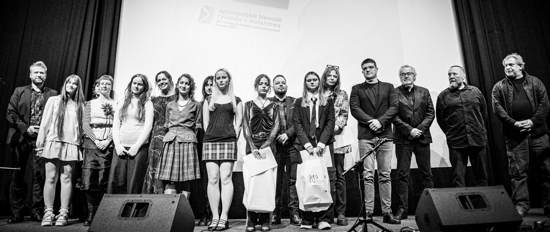 Czarno-białe zdjęcie przedstawia dużą grupę osób, w tym głównie młodych kobiet i mężczyzn oraz kilku starszych mężczyzn, stojących na scenie. Grupa jest ustawiona w linii, frontem do widza, i prawdopodobnie są to laureaci lub uczestnicy wystawy.
Elementy wizualne:
Tło: Za grupą znajduje się duży, jasny ekran projekcyjny z widocznym logotypem imprezy. Logo zawiera duży numer "17" oraz mniejszy tekst w języku polskim: "ogólnopolskie biennale rysunku i malarstwa". Tło ekranu jest jasne, otoczone ciemnymi kurtynami.
Osoby: Grupa liczy czternaście osób. Większość młodych osób stoi w środku i ma swobodny, zróżnicowany ubiór. Niektórzy trzymają w rękach teczki, dyplomy lub torby z nagrodami. Starsze osoby stoją głównie po bokach.
Pierwszy plan: Na pierwszym planie widać ciemne głośniki (monitory sceniczne) oraz kable i statywy mikrofonowe.
Oświetlenie jest mocne i skupione na grupie, kontrastując z ciemnym otoczeniem sceny.