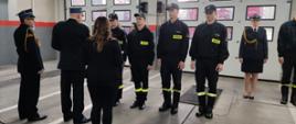 Awansowani strażacy stoją przed Komendantem i Zastępcą Komendanta. W ich tle strażacy osoby cywilne i bramy garażowe. 