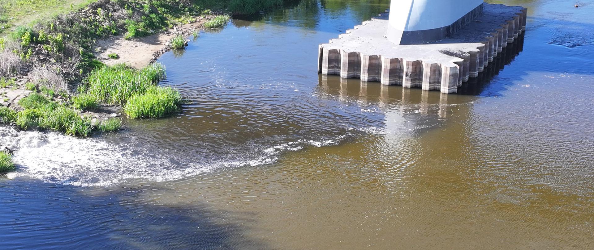 Powierzchnia rzeki o jasno – brązowym kolorze. Po lewej stronie widać brzeg rzeki oraz wpływające do niej spienione ścieki. Na górze widać białą podstawę przęsła mostu.