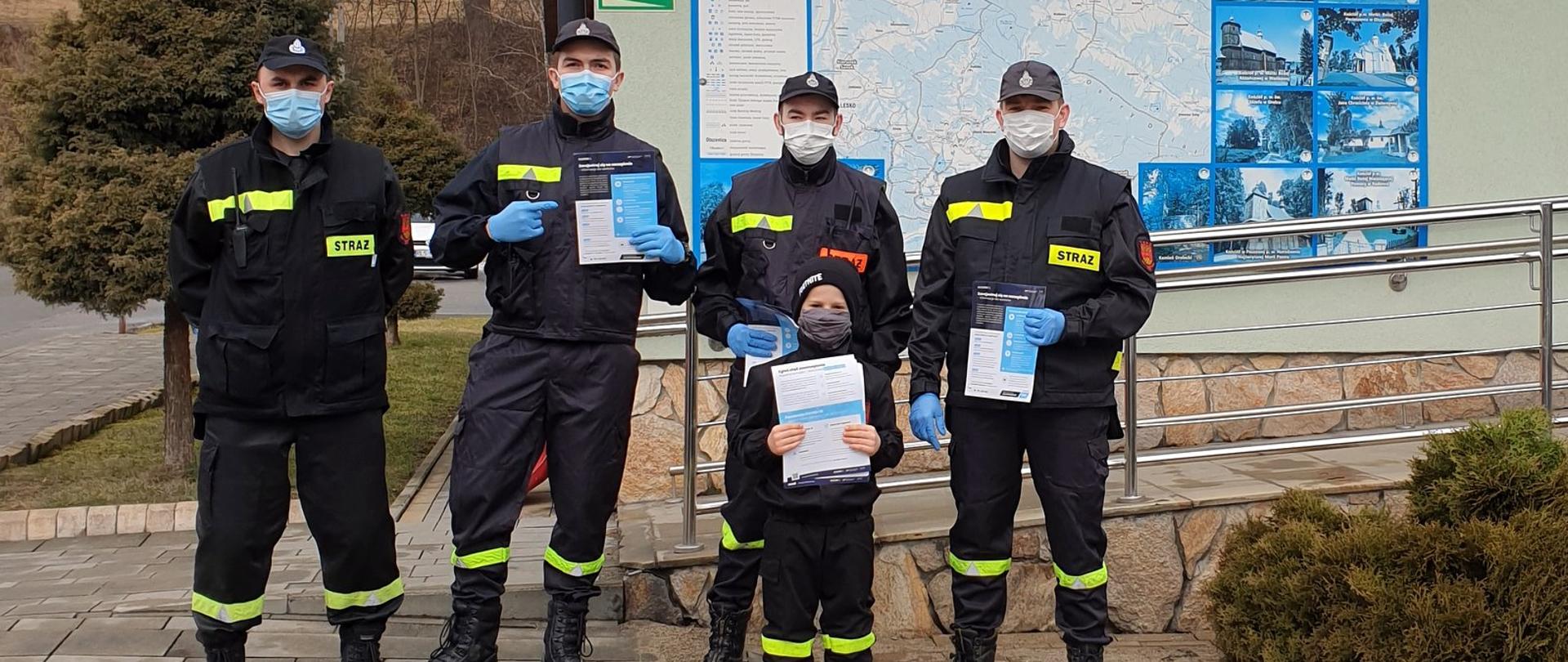 Na zdjęciu w rzędzie stoi czterech strażaków OSP z ulotkami w rękach. Przed nimi stoi mały chłopiec w mundurze OSP, także z ulotką w rękach. Za nimi tablica informacyjna z napisem gmina Olszanica.