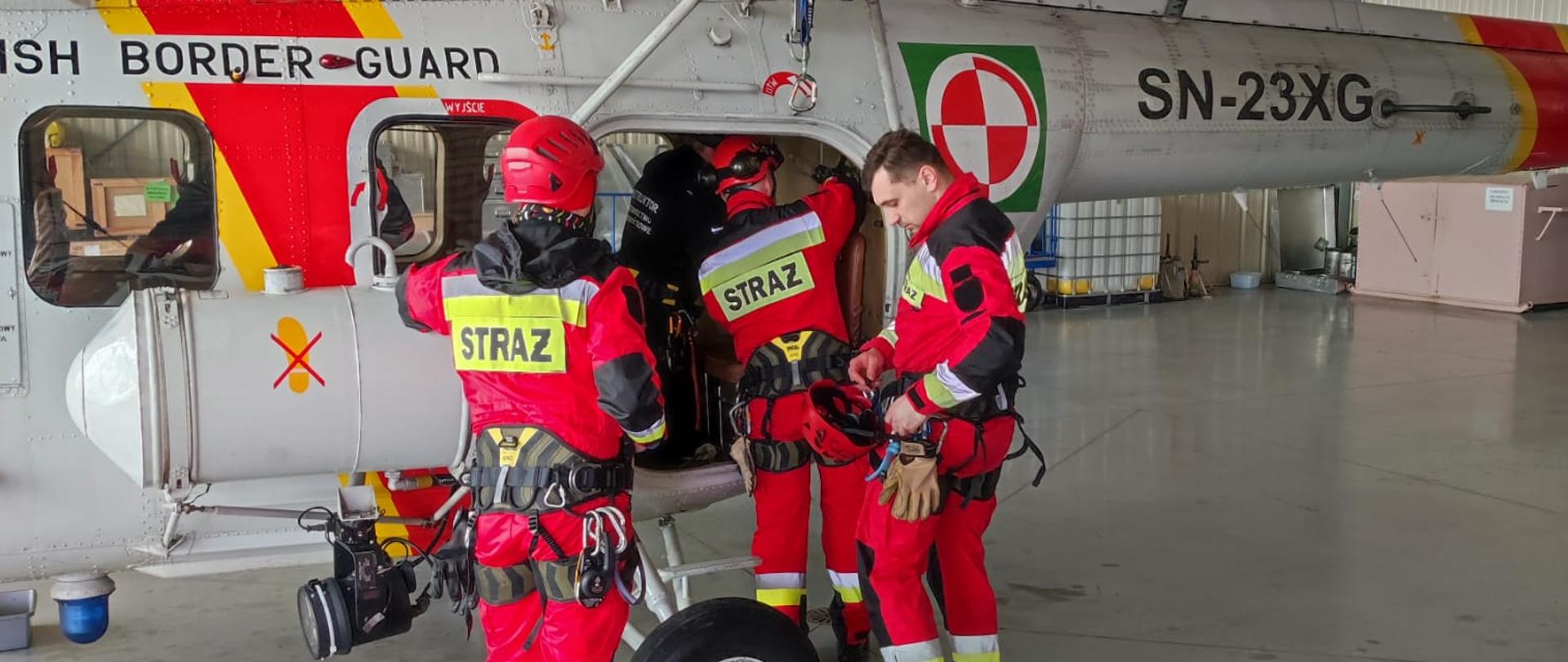 Na zdjęciu znajduje się śmigłowiec w garażu Straży Granicznej oraz strażacy wsiadający do śmigłowca.