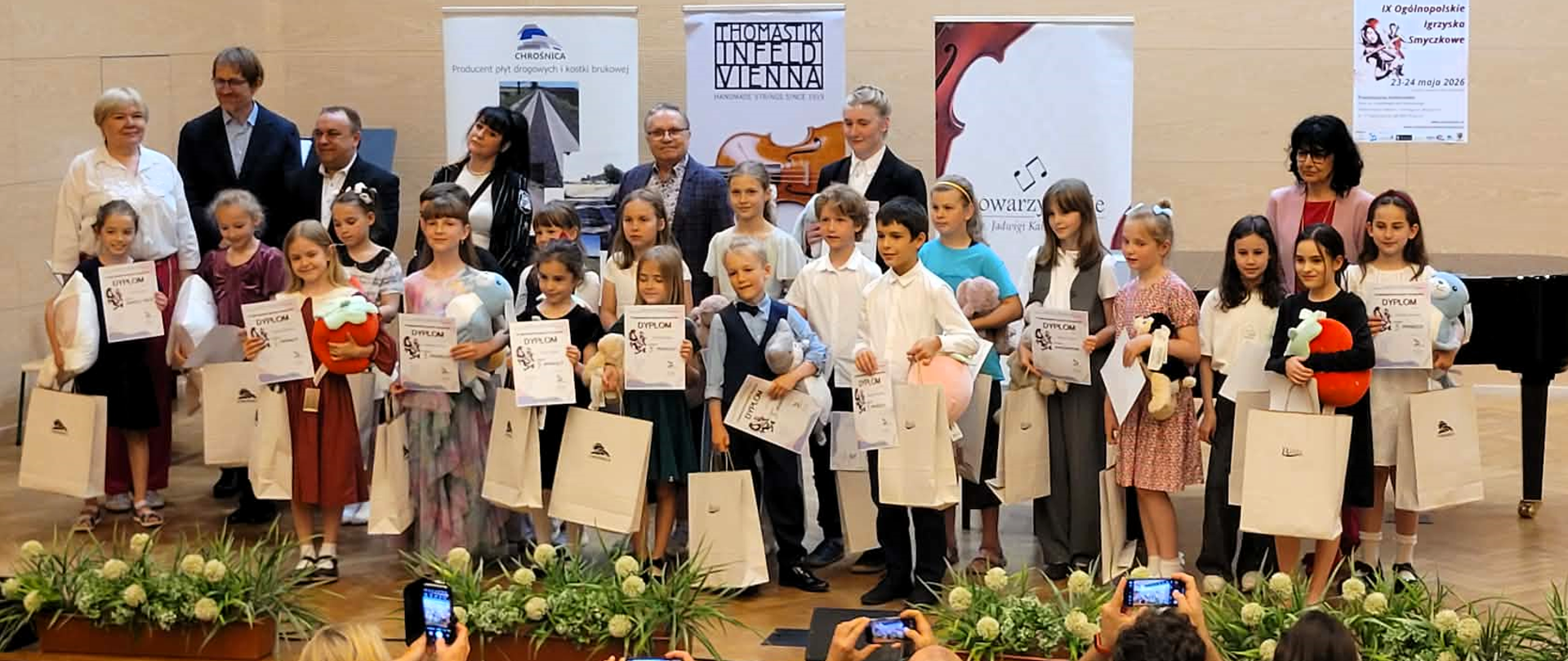 grupa kilkunastu dzieci z dyplomami stojących na scenie, za nimi banery konkursowe i kilku dorosłych. Przed sceną widać kilka kilka rzędów foteli na widowni.