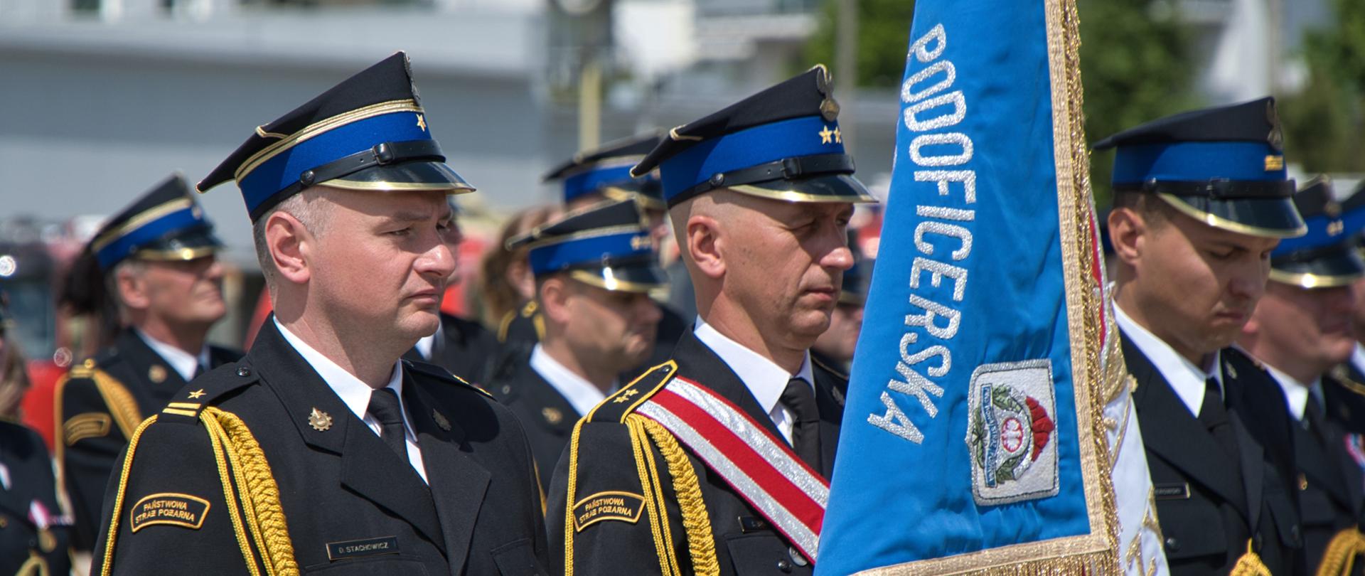 Poczet sztandarowy z 3 strażaków. Na pierwszym planie widać poczet - niebieskie tło z napisem podoficerska