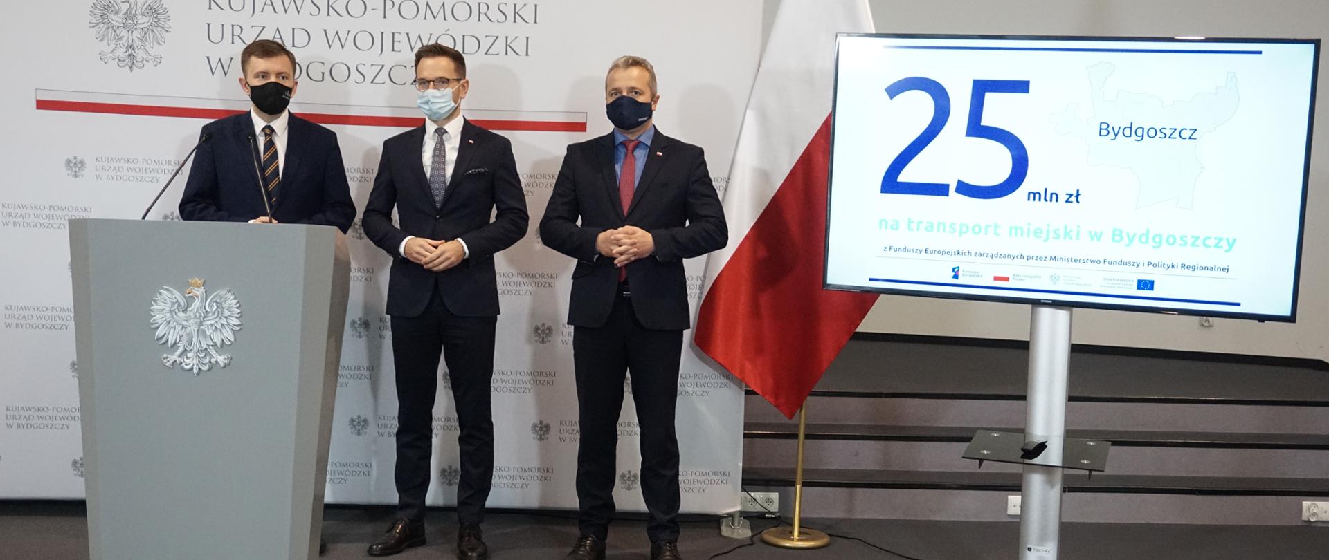 Waldemar Buda w mównicy. Za nim dwóch mężczyzn. Obok ekran z grafiką, flaga i ścianka.