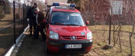 Zdjęcie przedstawia posesję prywatną. Strażak Ochotniczej Straży Pożarnej w Rakszawie asystuje mężczyźnie w starszym wieku w trakcie wysiadania z samochodu pożarniczego po powrocie ze szczepienia.