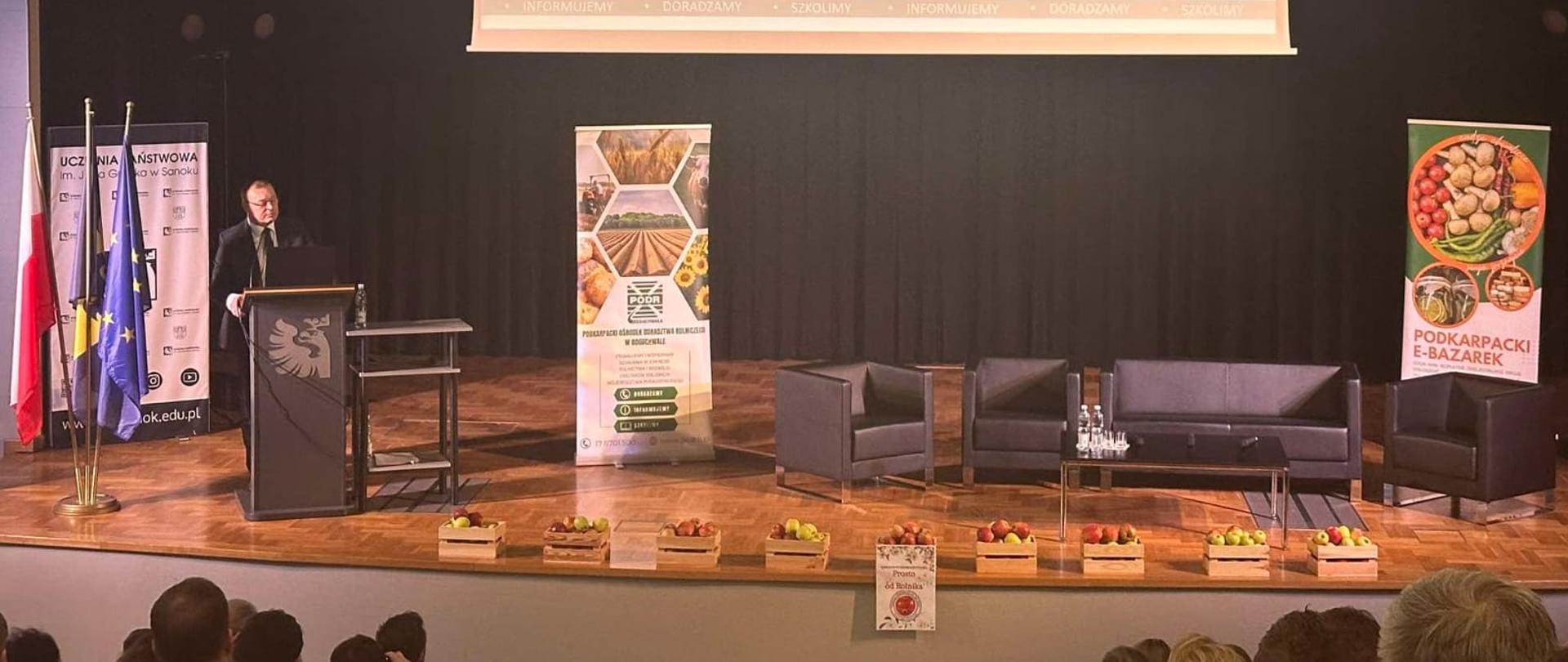 Scena z czarnym tłem. Po lewej stronie przy mównicy stoi mężczyzna w garniturze. Z przodu sceny skrzynki z jabłkami, z sufitu spuszczony ekran z prezentacją.