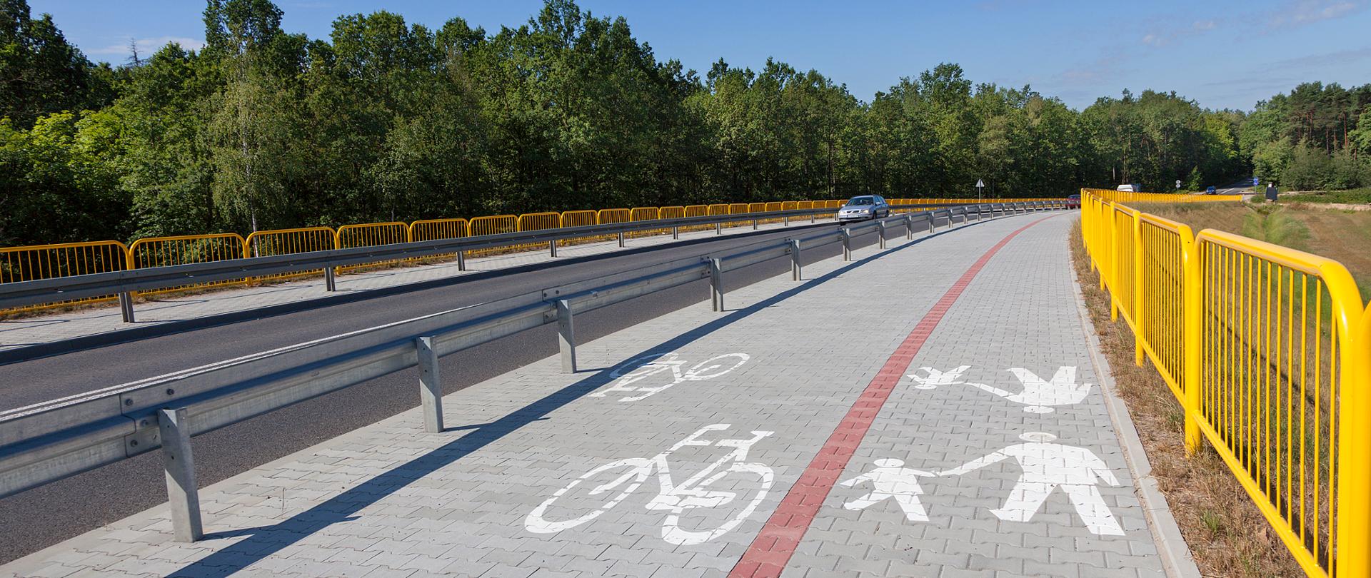 Fotografia zintegrowanej ścieżki rowerowej z chodnikiem biegnąca wzdłuż drogi. Na pierwszym planie chodnika i ścieżki rowerowej widoczne oznakowanie poziome. Droga oddzielona od ścieżki barierami ochronnymi, a całość zabezpieczona barierami ochronnymi w kolorze żółtym. Na drodze poruszają się pojazdy osobowe. 