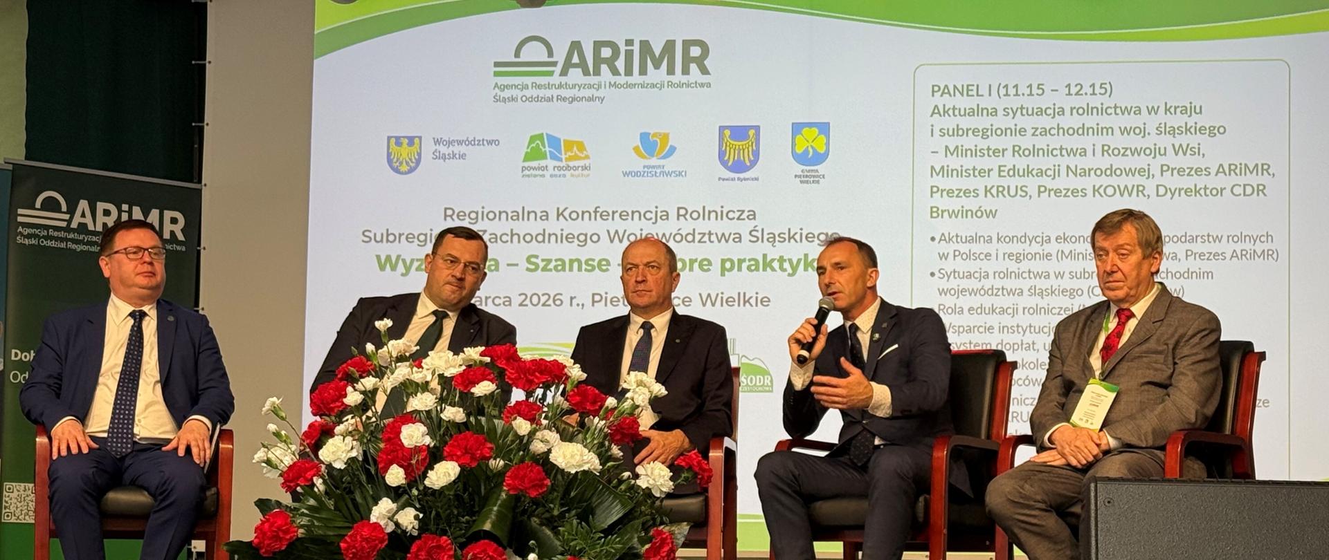 Pięciu mężczyzn w garniturach siedzi na fotelach na scenie. Jeden z nich Prezes KRUS trzyma mikrofon. Za nimi na dużym ekranie wyświetlana jest prezentacja z logo ARiMR. Przed mężczyznami na scenie stoi duży kosz białych i czerwonych goździków.