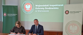 Przy stole na sali konferencyjnej siedzą Mazowiecki Wojewódzki Inspektor Ochrony Środowiska Krzysztof Cząstkiewicz i Mazowiecki Wojewódzki Lekarz Weterynarii Izabela Floryszczyk. Pani Floryszczyk składa podpis na swojej kopii porozumienia, Pan Cząstekiwicz patrzy się w obiektyw aparatu.