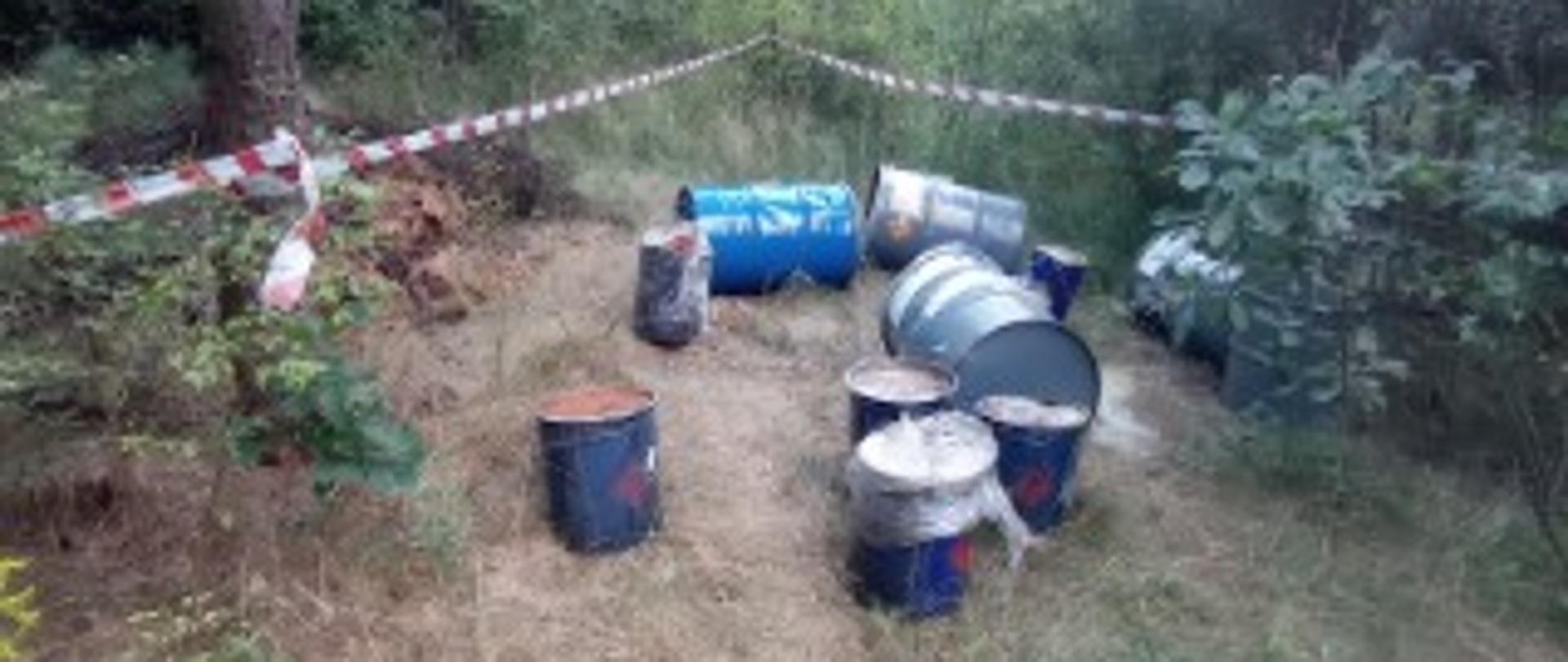 Beczki o pojemności 200 l i wiadra o pojemności 30 l leżą porozrzucane na odsłoniętym leśnym terenie.