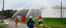 Strażak rozmawia z pracownikiem Bazy Paliw. Rozmowa jest na tle zbiornika na który podawana jest woda w trakcie ćwiczeń z 2 działek. Strażacy podający wodę na zbiorniki urania są w ubrania żaroochronne. W tle widać 2 samochody pożarnicze.