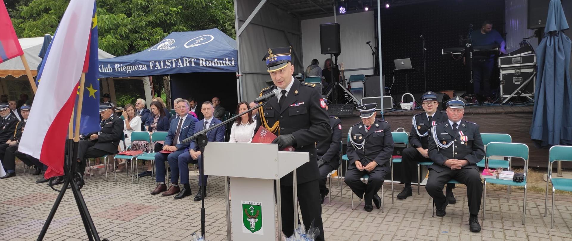 na zdjęciu st. bryg. Dariusz Gieroń - Zastępca Opolskiego Komendanta Wojewódzkiego Państwowej Straży Pożarnej podczas przemawiania, w tle pozostali zaproszeni goście oraz scena. Obok mównicy Flaga Polski oraz Flaga Unii Europejskiej.