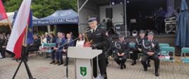 na zdjęciu st. bryg. Dariusz Gieroń - Zastępca Opolskiego Komendanta Wojewódzkiego Państwowej Straży Pożarnej podczas przemawiania, w tle pozostali zaproszeni goście oraz scena. Obok mównicy Flaga Polski oraz Flaga Unii Europejskiej.