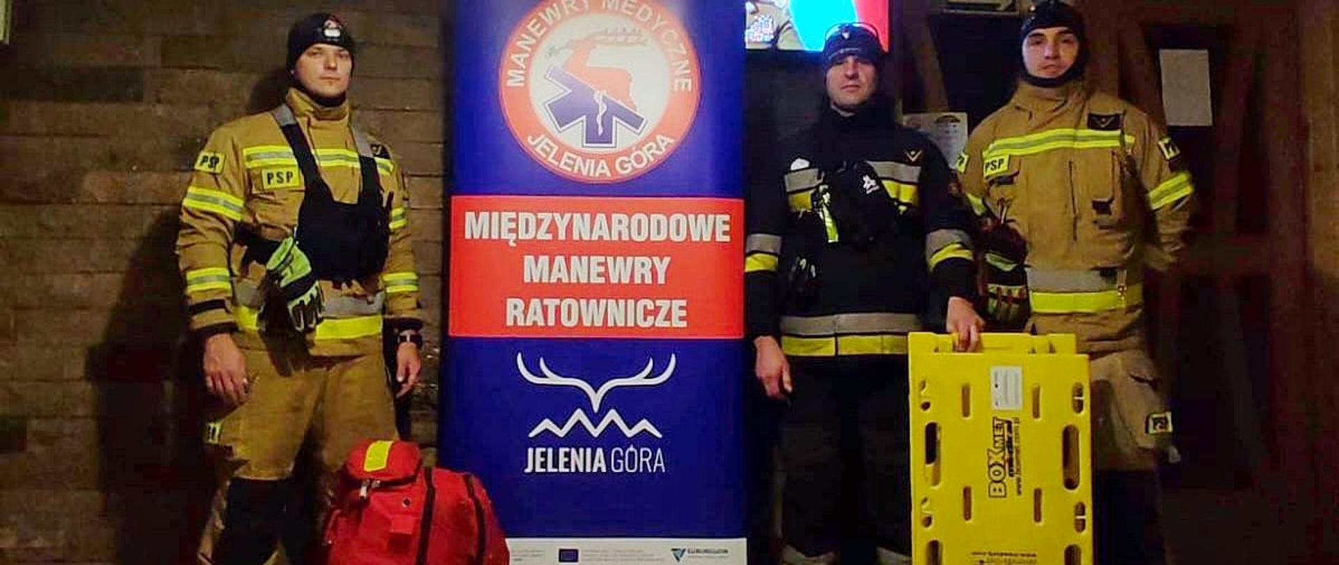 Zdjęcie przedstawia trzech funkcjonariuszy KM PSP w Sosnowcu oraz rolap z grafiką oraz napisem „Międzynarodowe manewry ratownicze Jelenia Góra”
