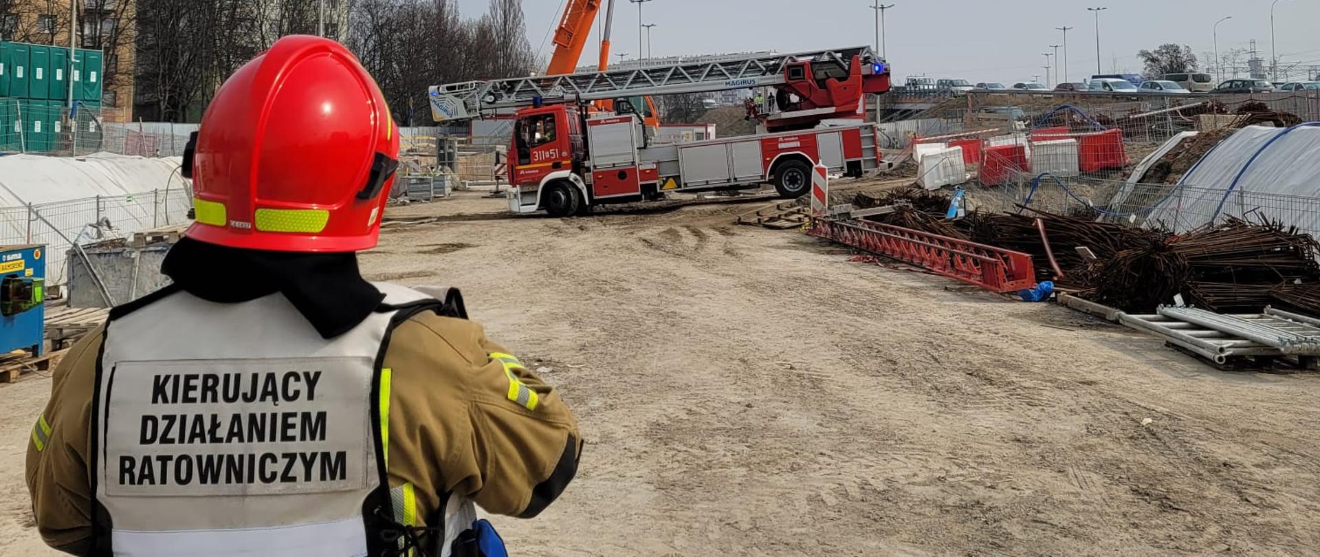 Strażak na terenie budowy w kamizelce z napisem kierujący działaniem ratowniczym, w tle drabina mechaniczna.