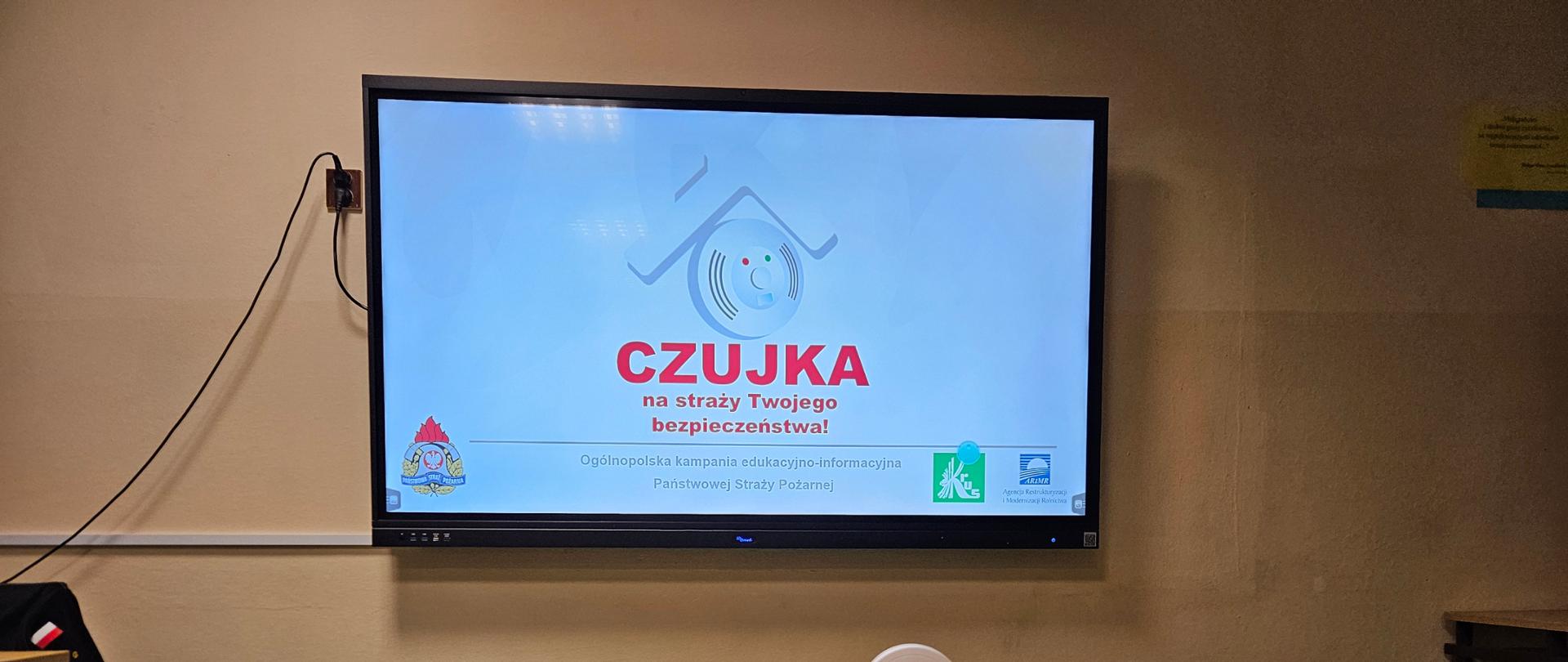 Ekran prezentuje slajd informacyjny z hasłem: "CZUJKA na straży Twojego bezpieczeństwa!" (Wykrywacz/Czujnik czuwa nad Twoim bezpieczeństwem!). Poniżej widnieje napis: "Ogólnopolska kampania edukacyjno-informacyjna Państwowej Straży Pożarnej" (Ogólnopolska kampania edukacyjno-informacyjna Państwowej Straży Pożarnej). Na drewnianym stole przed ekranem ustawione są cztery różne urządzenia przypominające czujniki dymu lub tlenku węgla, w kolorach białym i szarym, służące jako rekwizyty.