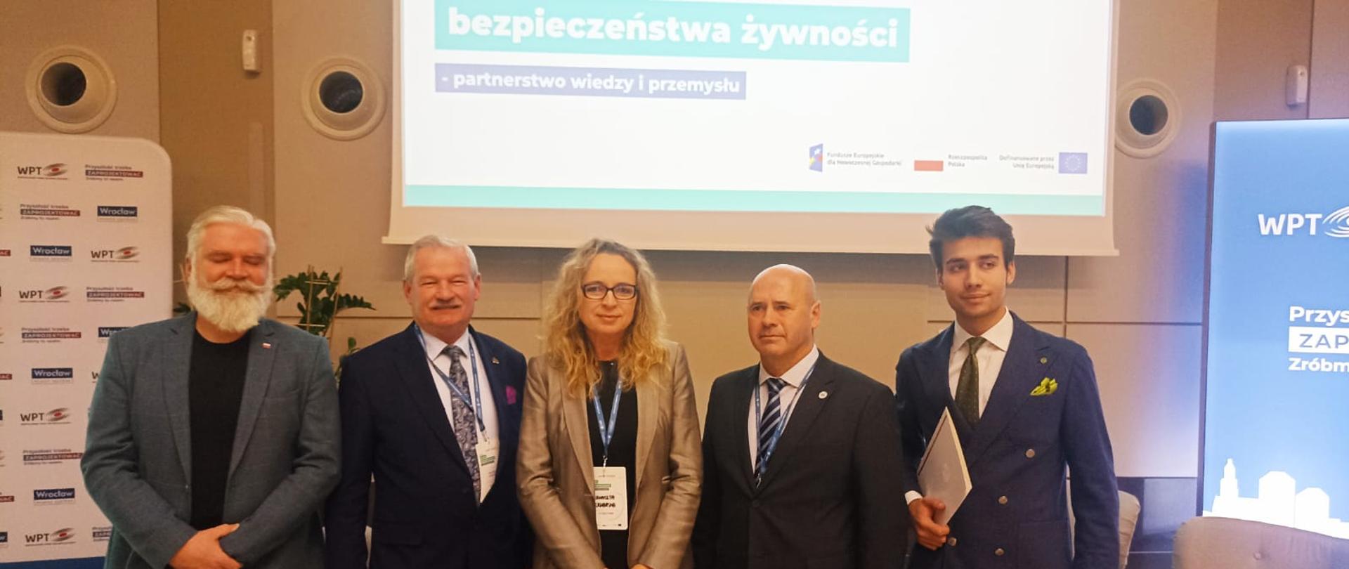 
Uczestnicy Konferencji stojący przy ekranie projekcyjnym.