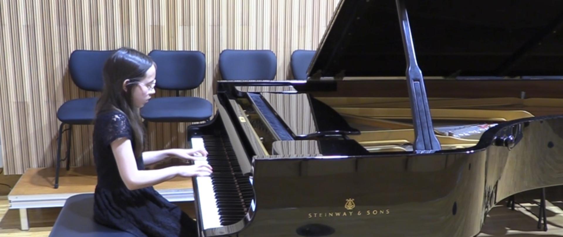 Dziewczynka w czarnej, koronkowej sukience, z długimi, ciemnymi włosami i w okularach, gra na fortepianie.