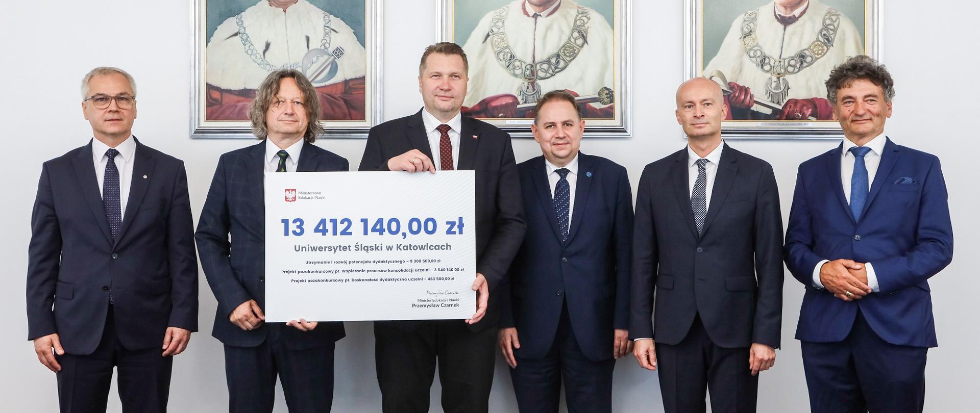 Minister Czarnek i pięciu mężczyzn w garniturach stoją pod ścianą na której wiszą 3 portrety, dwóch mężczyzn trzyma symboliczny czek z napisem 13 412 140 zł.
