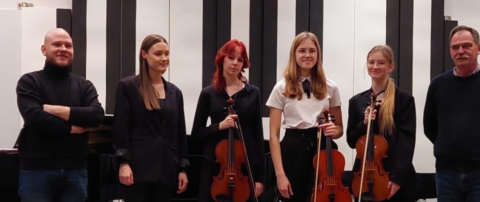 W jednym rzędzie stoją trzy nastolatki trzymające przed sobą w rękach altówki i kobieta, po bokach stoi dwóch mężczyzn.