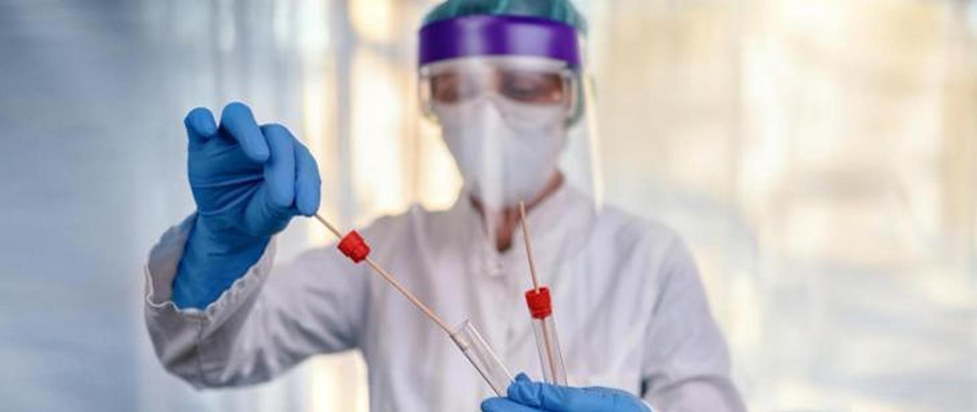 Pracownik laboratorium w środkach ochrony osobistej pobiera próbki do badań laboratoryjnych przy użyciu wymazówek i probówek. Zdjęcie symbolizuje diagnostykę, bezpieczeństwo zdrowotne oraz pracę laboratoryjną w obszarze zdrowia publicznego.
