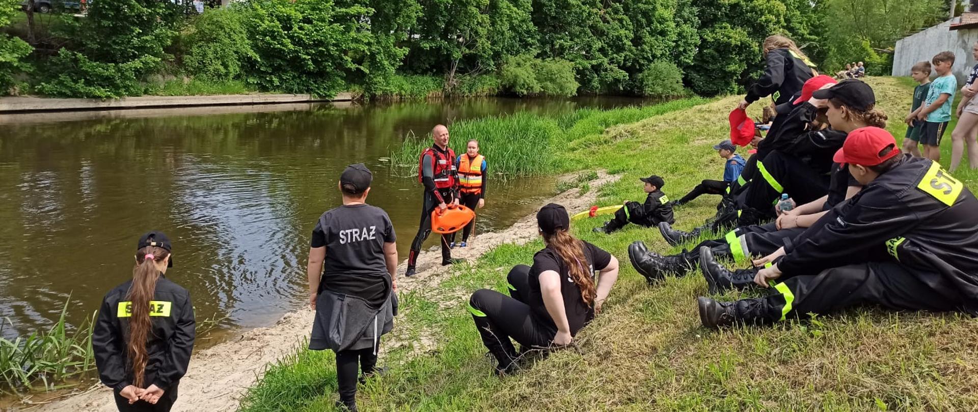 Nad brzegiem rzeki siedzą dzieci w mundurkach strażackich, obserwują instruktora który stoi przy rzece.
