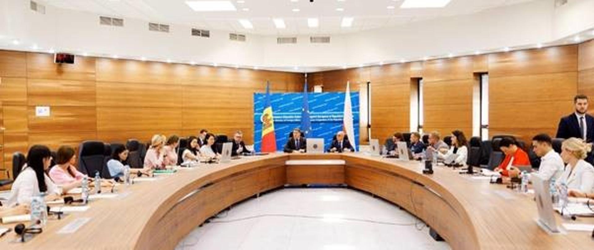 Nowoczesna sala konferencyjna. Przy dużym drewnianym zaokrąglonym stole siedzi kilkanaście osób, uczestników szkolenia. W tle widoczne są flagi Mołdawii, Unii Europejskiej i Polski.
